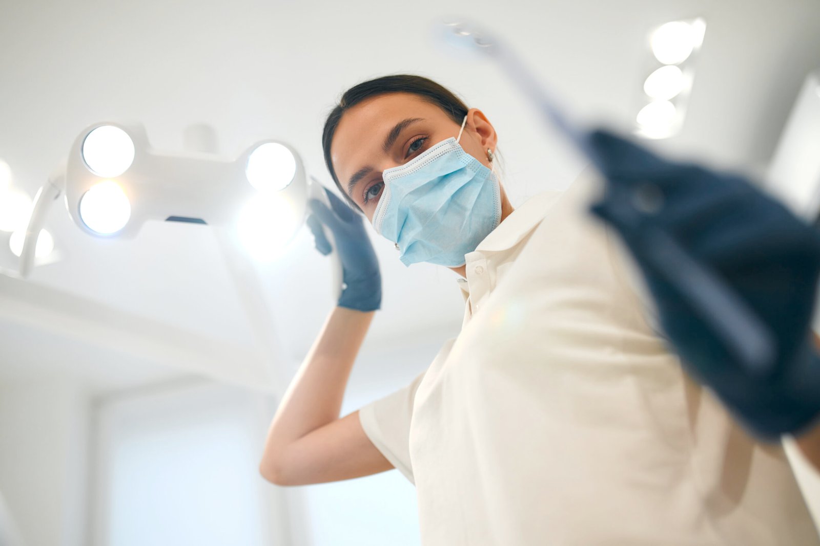 Bottom view of a young female dentist in a white uniform and medical mask in a dental office with modern equipment, holding a lamp and tools.